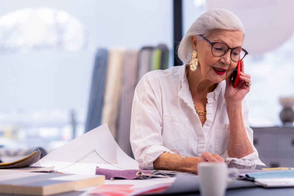 Telephone conversation. Enchanting well-groomed aging boss in white stylish shirt and trendy accessories busily speaking on the phone in office.